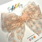A blush-colored sheer lace bow with embroidered snowflake details, displayed on a white card with the brand name 'Parker & Kay' printed on it.