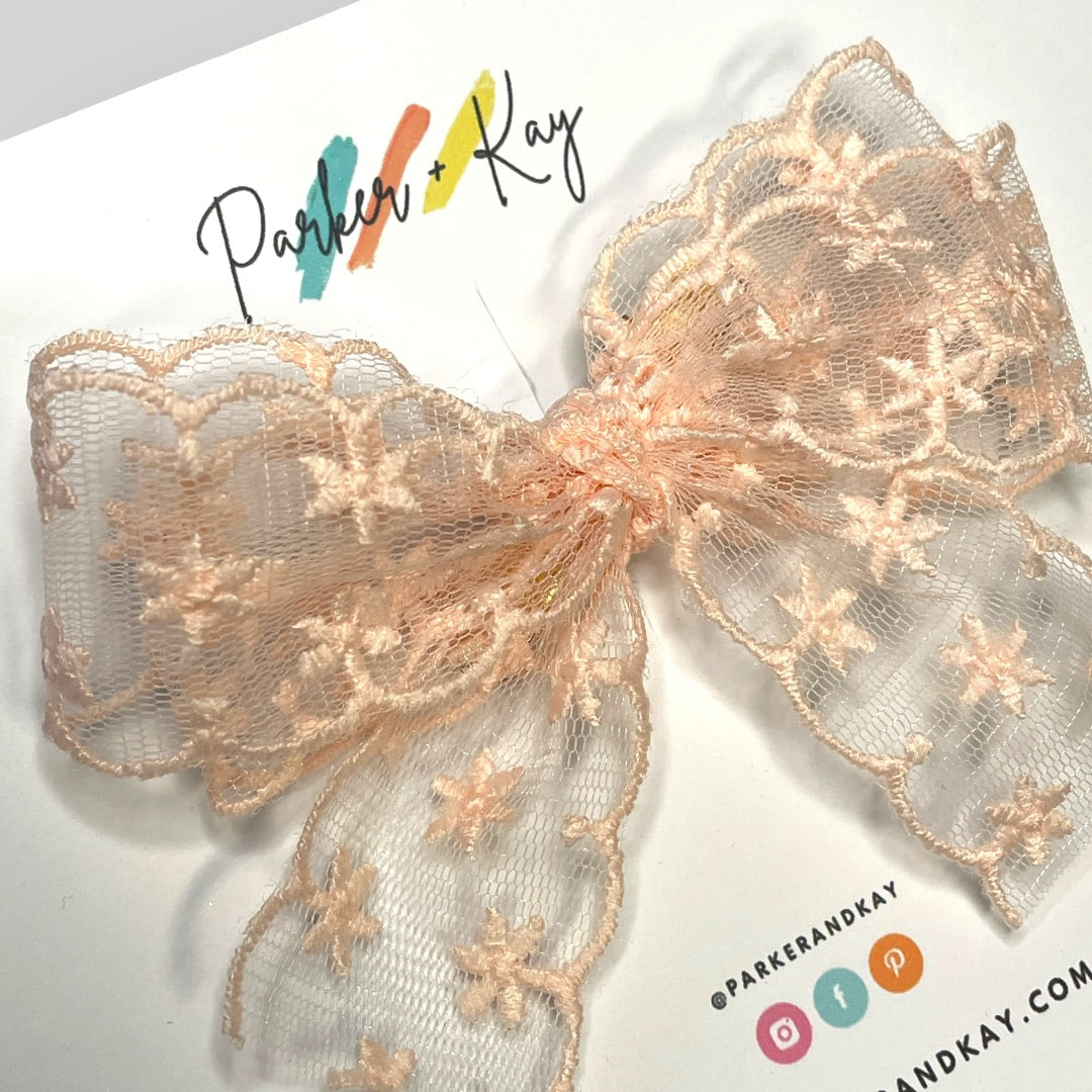 A blush-colored sheer lace bow with embroidered snowflake details, displayed on a white card with the brand name 'Parker & Kay' printed on it.