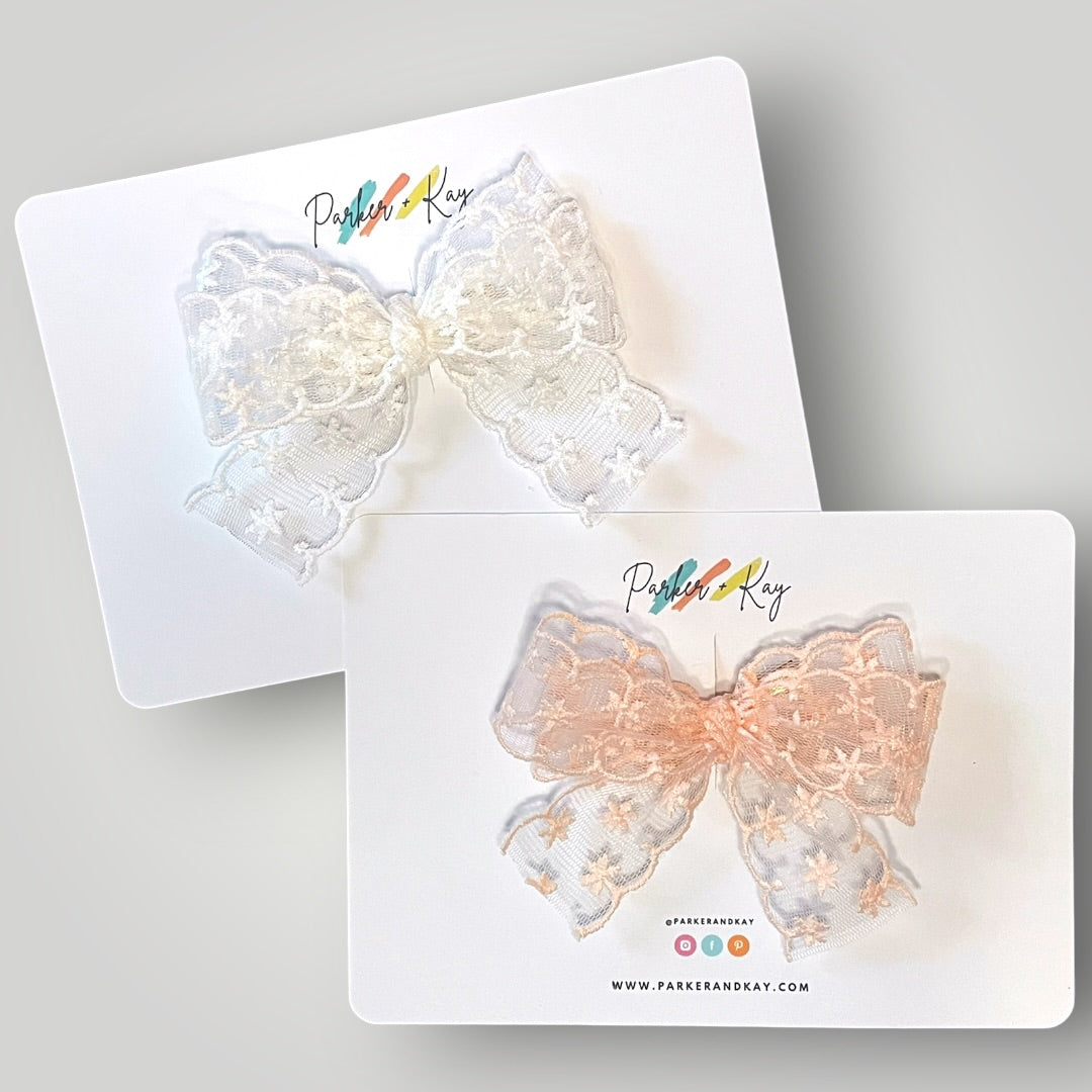 Two decorative bows on white display cards against a light gray background. The top bow is white lace with small embroidered snowflakes.  The bottom bow is blush lace with small embroidered snowflakes.
