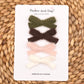 Four cashmere hair bows in olive green, dark brown, light pink, and cream white, arranged vertically on a white display card from Parker and Kay Collective, placed on a woven straw mat background.