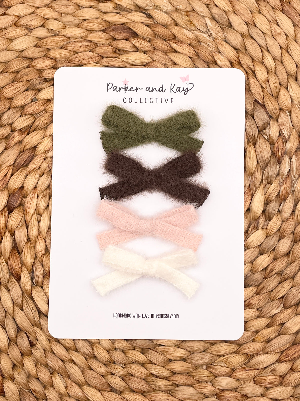 Four cashmere hair bows in olive green, dark brown, light pink, and cream white, arranged vertically on a white display card from Parker and Kay Collective, placed on a woven straw mat background.