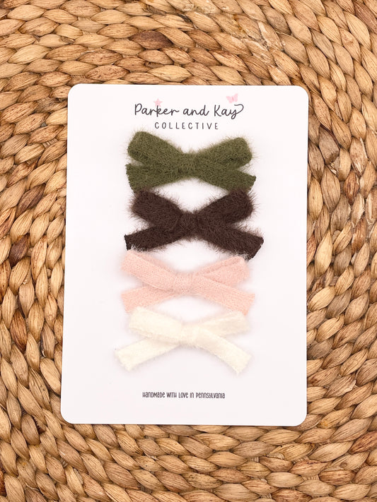 Four cashmere hair bows in olive green, dark brown, light pink, and cream white, arranged vertically on a white display card from Parker and Kay Collective, placed on a woven straw mat background.
