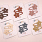 Six pairs of flower-shaped hair clips in different colors, each mounted on a white display card. The clips feature fabric flower petals with small pearl-like center details and are arranged on a light pink background.