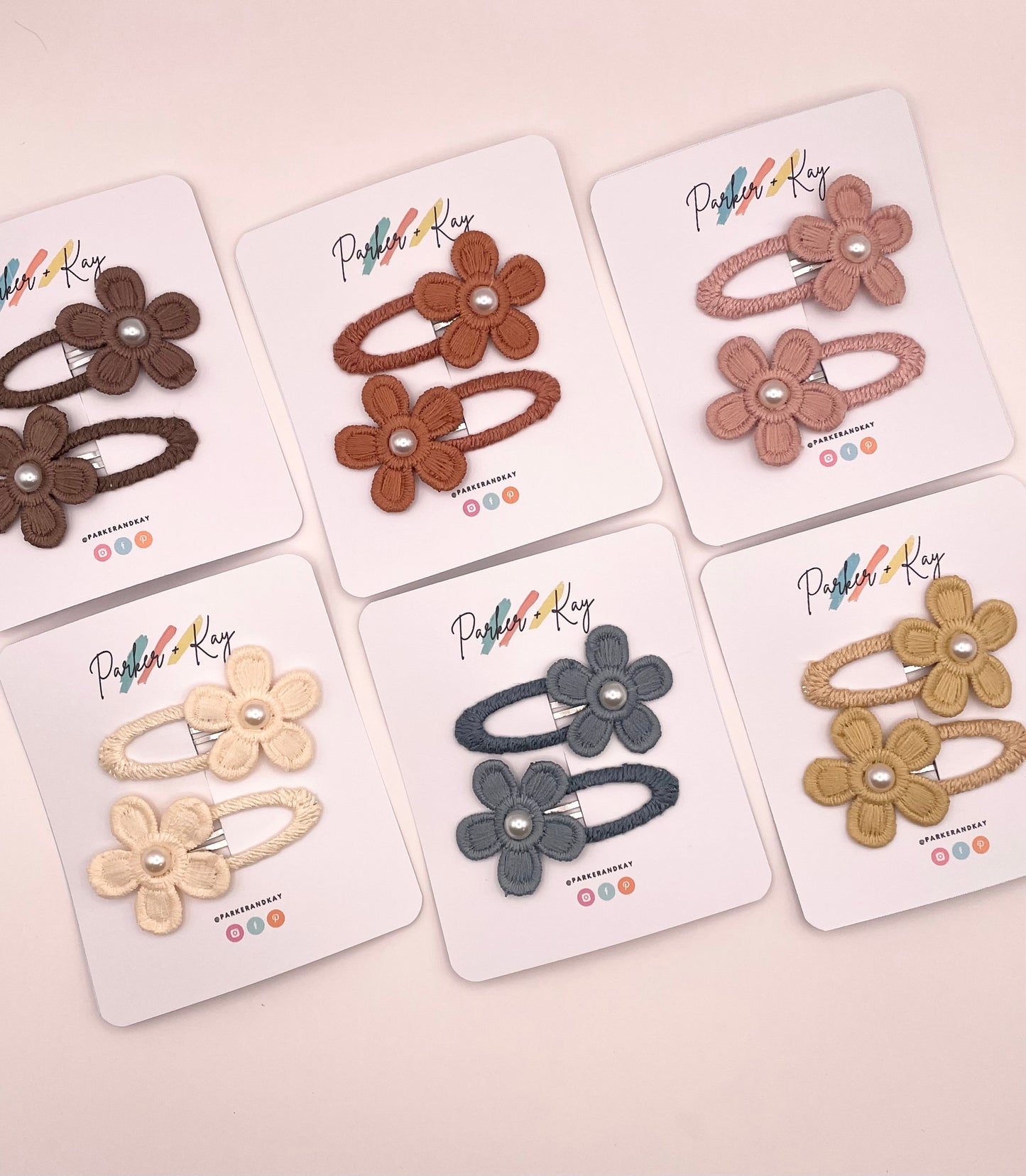 Six pairs of flower-shaped hair clips in different colors, each mounted on a white display card. The clips feature fabric flower petals with small pearl-like center details and are arranged on a light pink background.