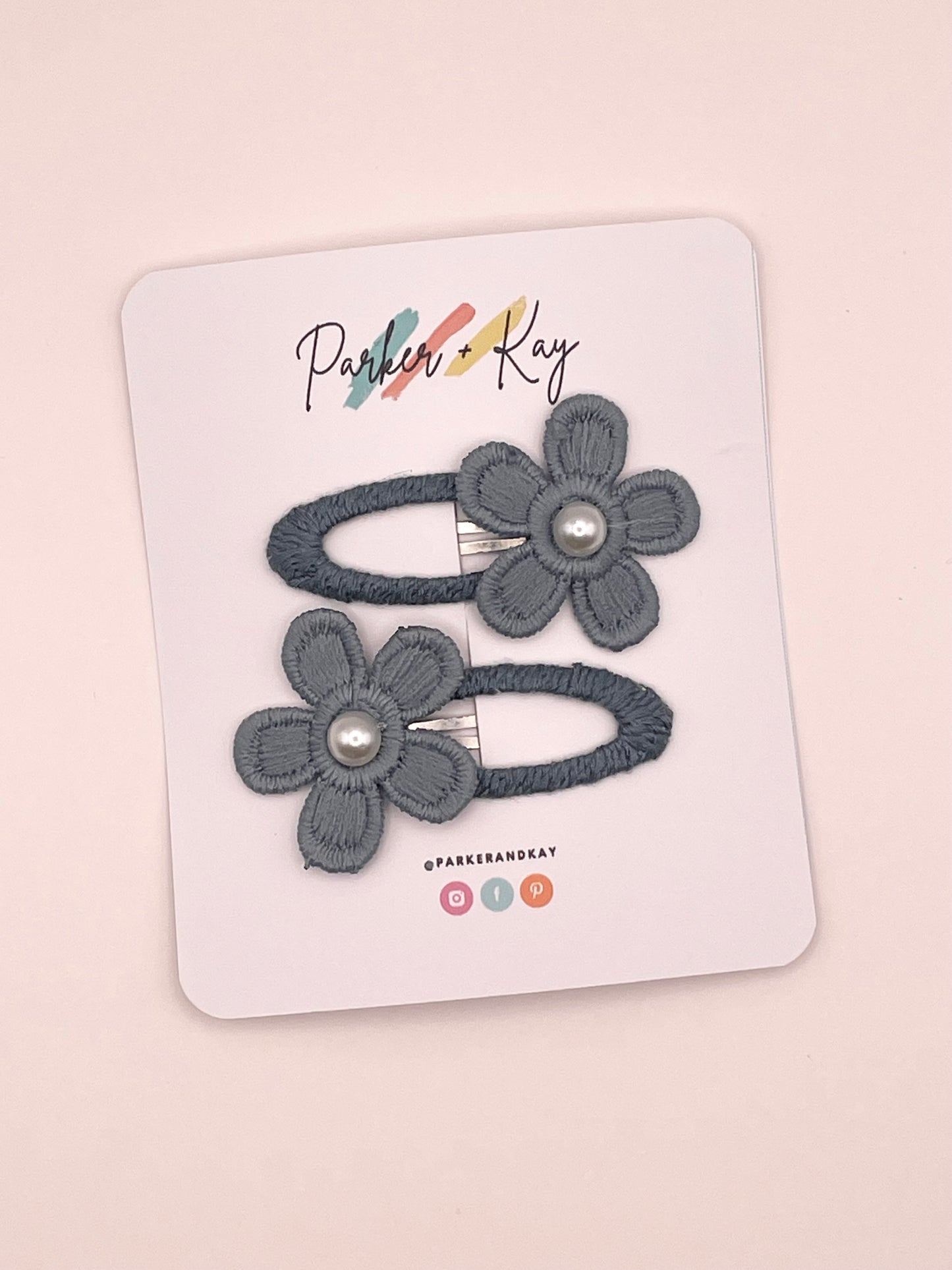 Two bluish-gray fabric flower hair clips with pearl centers, attached to a white display card printed with the brand name 'Parker & Kay'.