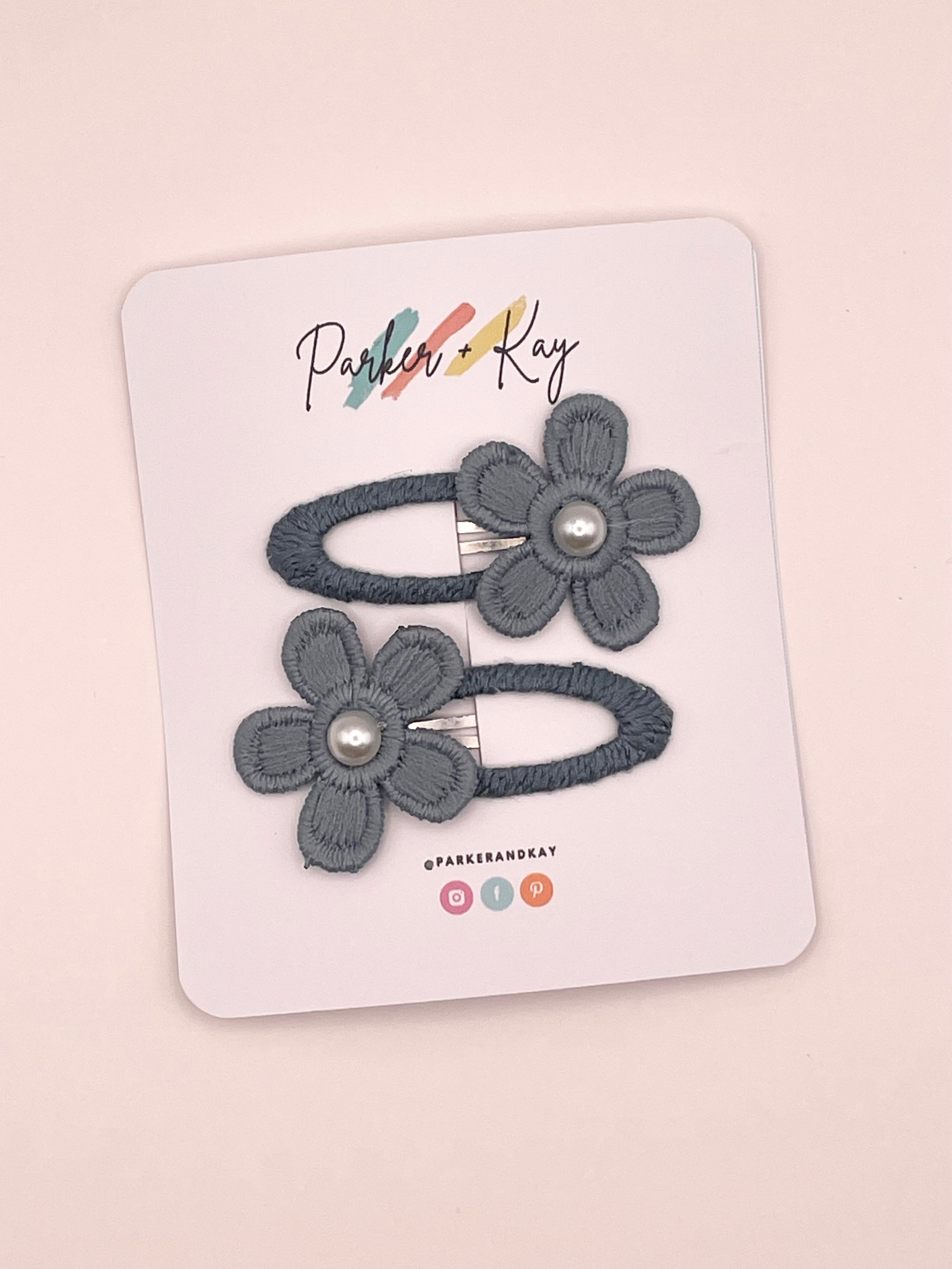 Two bluish-gray fabric flower hair clips with pearl centers, attached to a white display card printed with the brand name 'Parker & Kay'.