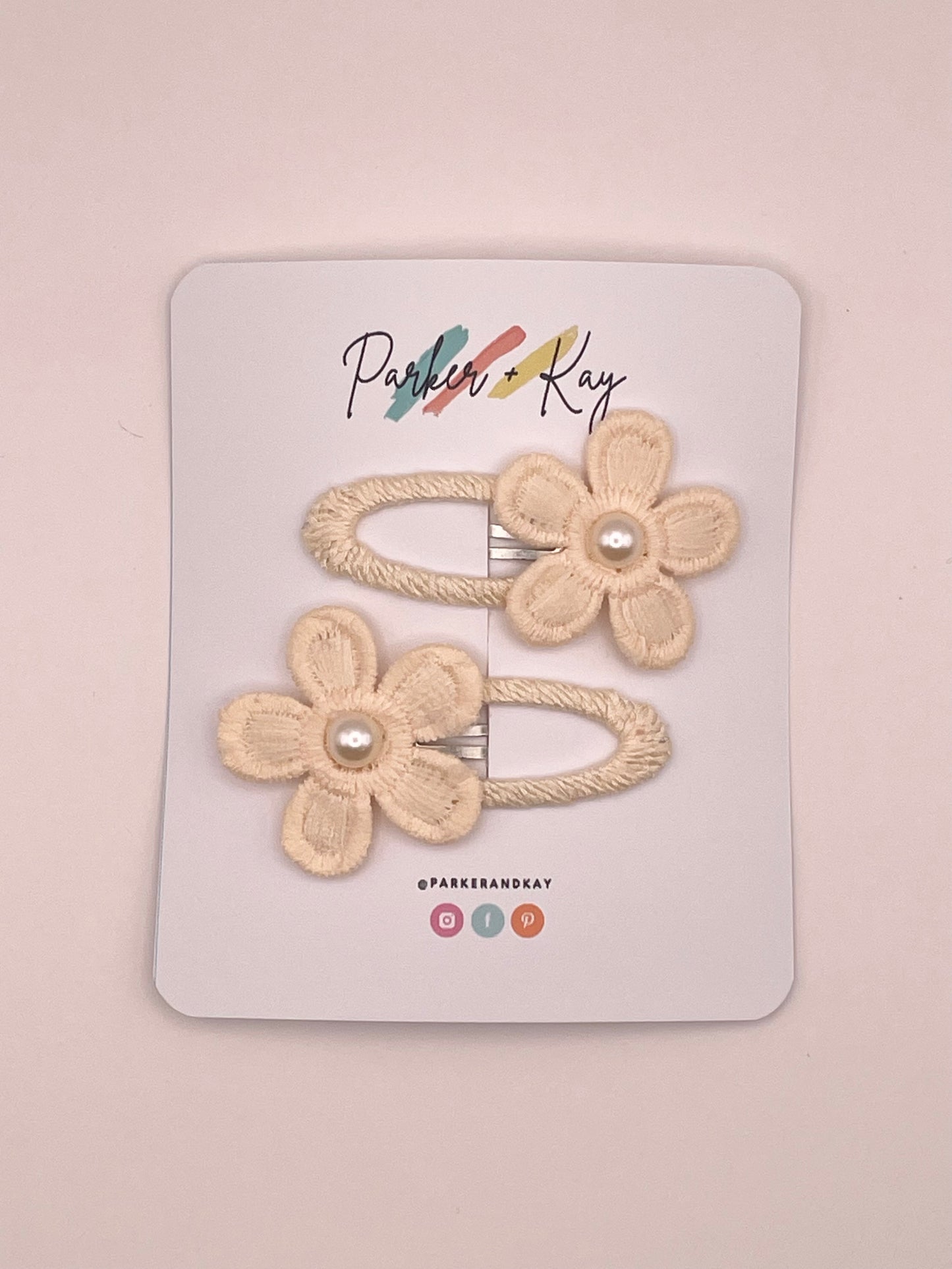 Two cream-colored fabric flower hair clips with pearl centers, attached to a white display card branded 'Parker + Kay'.