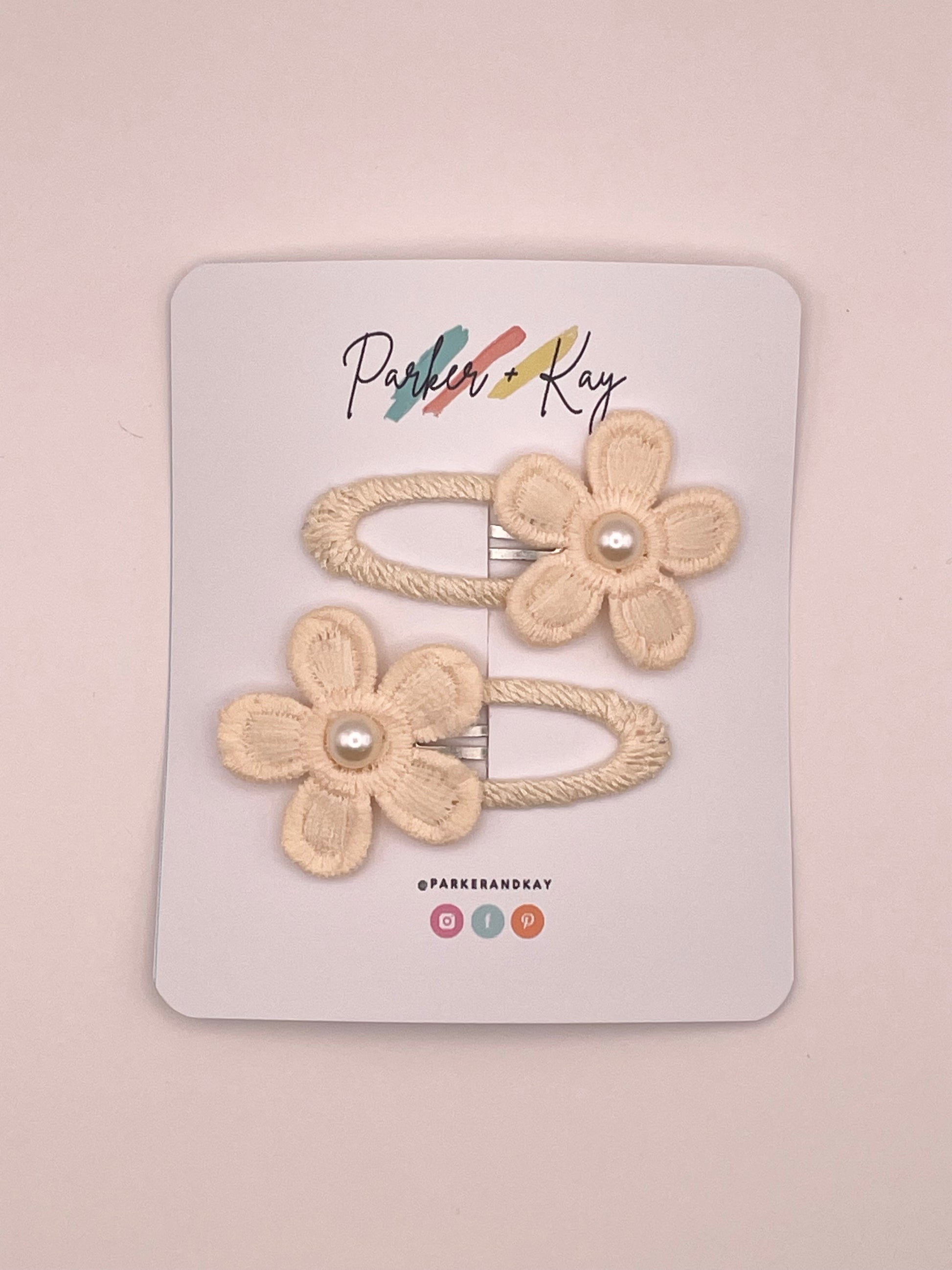 Two cream-colored fabric flower hair clips with pearl centers, attached to a white display card branded 'Parker + Kay'.